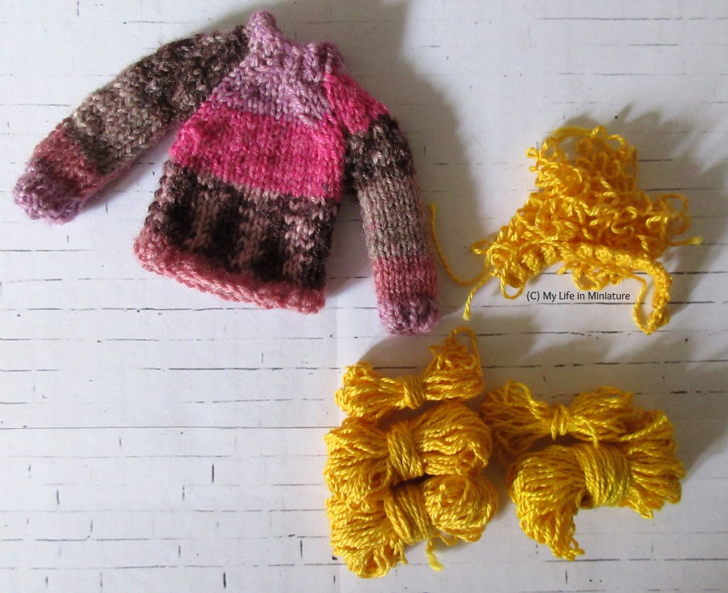 Petra's pink jumper sits on a white brick background. Next to it are five small skeins of wrinkled yellow yarn, and a small pile of the same yarn. 