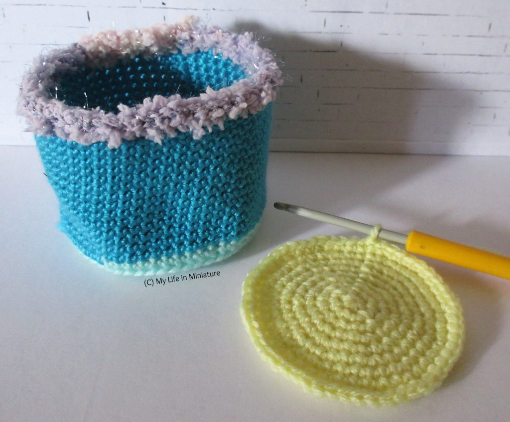 A blue crocheted pot with a purple fluffy rim sits against a white background. Beside it is a flat circle of yellow crochet, hook in the working loop.