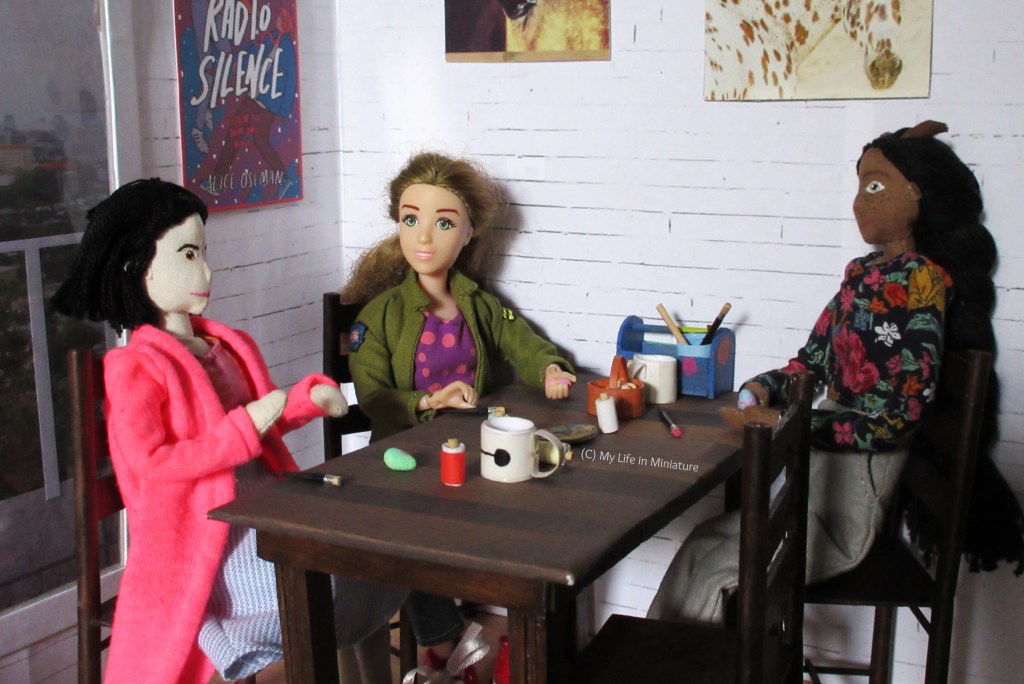 Sarah, Tiffany and Petra sit around Sarah's dining room table. Tiffany is gesturing, explaining an egg design to Sarah and Petra, who are paused mid-painting and listening intently. 