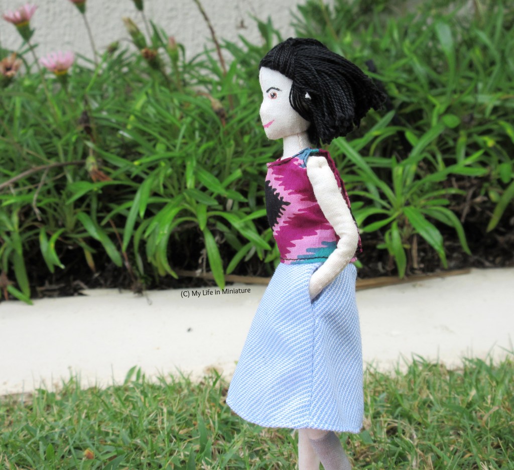 Tiffany stands on grass, next to a flower bed. She wears the tie-dyed shirt with a blue-and-white-striped skirt, hands in pockets. She faces the left, and her hair is blown in the wind behind her.