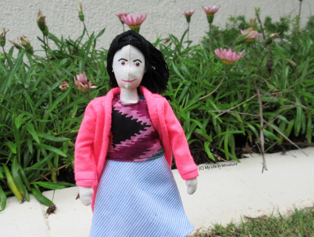 Tiffany stands on grass, in front of a flower bed. She wears the tie-dyed shirt and blue skirt, with her pink cardigan over the top. She faces the camera, hair being windblown to the right.