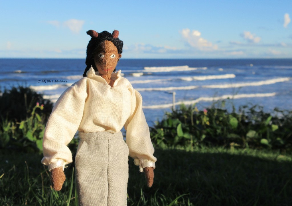 Petra stands on a grassy, vegetated hill overlooking the sea. Waves crash behind her. She wears the 'pirate' shirt and is smiling at the camera, hands by her sides.