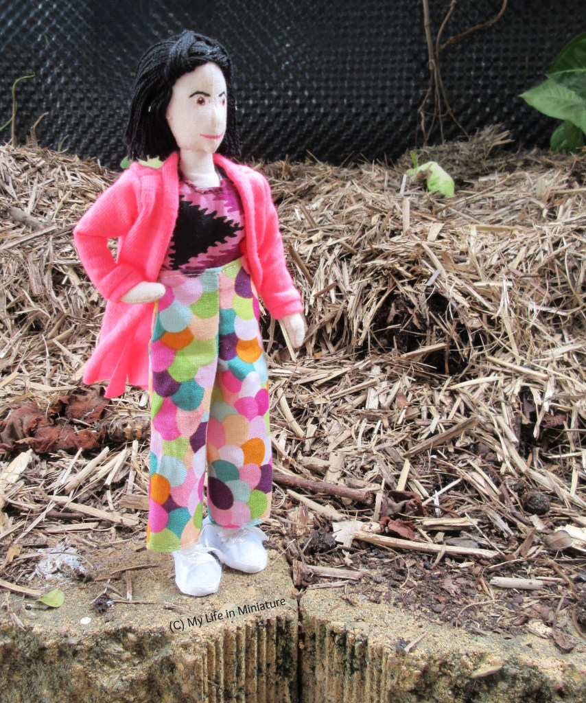 Tiffany stands on a brick on the edge of a mulched garden bed. She wears her multi-coloured pants with a purple tie-dyed shirt and a hot pink cardigan. She is placing one hand on her hip, and is about to walk to the right, looking in that direction.