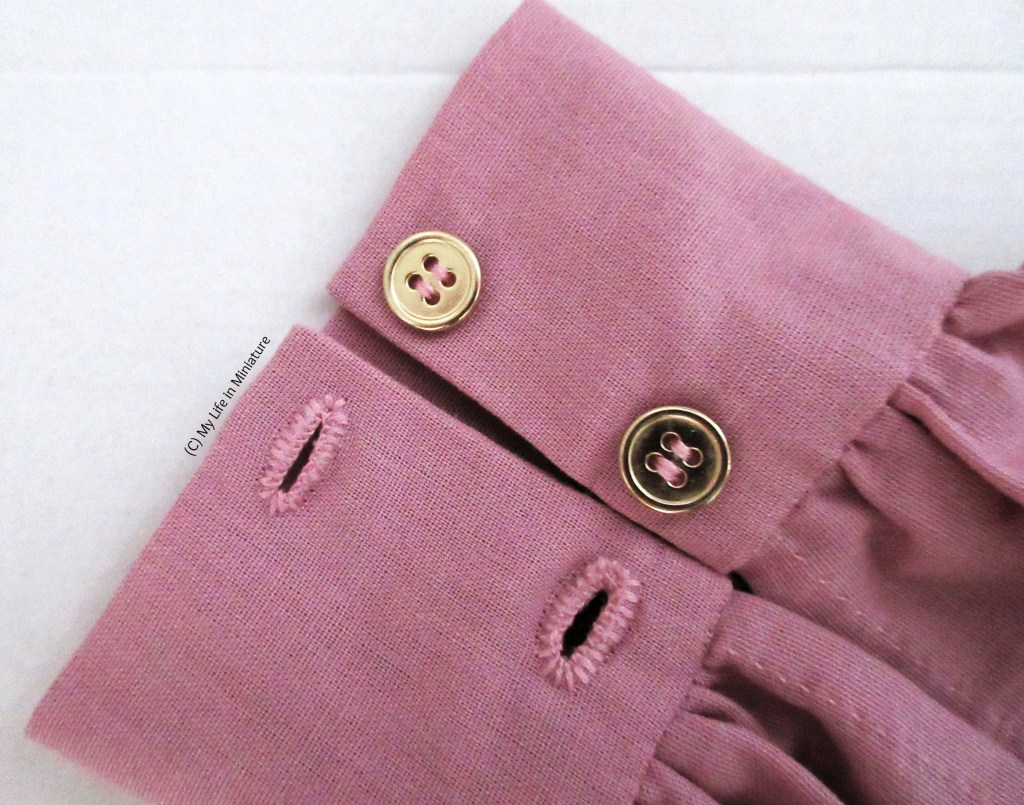 Close-up of the buttons and buttonholes of the cuff. Two hand-sewn buttonholes are on the opposite cuff edge to the gold buttons. The holes are sewn in a slightly paler pink than the fabric. 