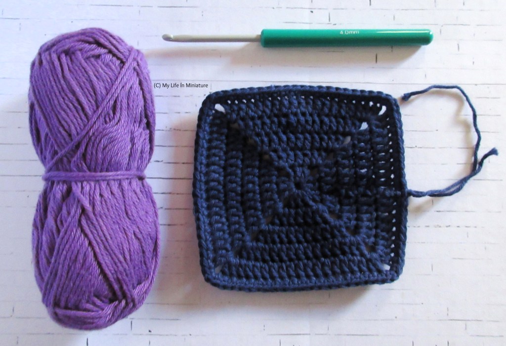 A solid navy crocheted square sits on a white brick background, ends visible. Above it is a 4mm crochet hook, and to its left is a ball of light purple cotton yarn. 