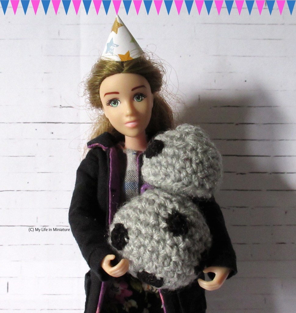 Sarah holds her android, Chip, in her arms and smiles at the camera. She wears a conical party hat on her head, and a string of triangular bunting is overlaid onto the top edge of the image. 