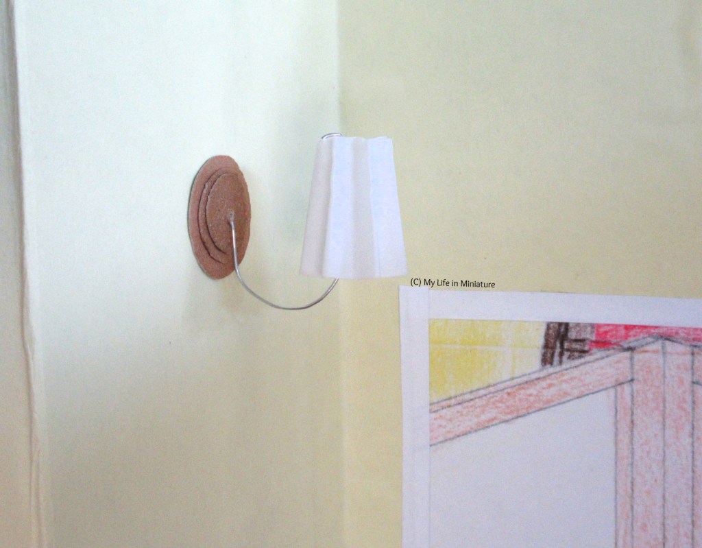 Close-up of one of the lights in the last image. They have a white concertina-d lampshade, attached to the wall with a smile-shaped loop of silver wire. The wall fitting is three brown circles, stacked smallest to largest. The corner of the doorway is visible in the bottom right.