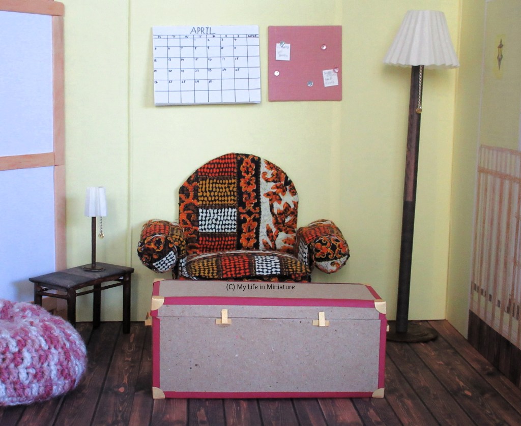 Image shows the entire upstairs room in the Palace Library. The door is on the left, the room divider is on the right, and the armchair and trunk are in the centre. Beside the door is a tall freestanding light with a white lampshade, and on the side table beside the armchair is a smaller version of it. 