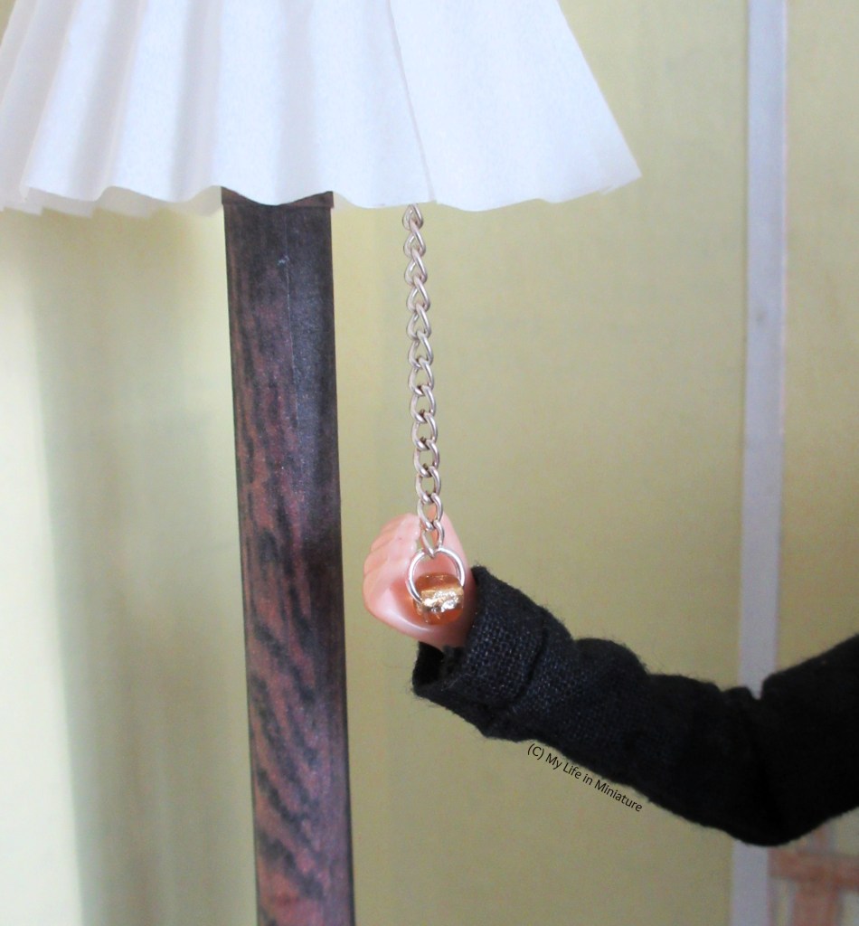 Close-up of the chain hanging from beneath the lampshade of the floor lamp. Sarah's hand cups the gold bead on the end of the chain. 