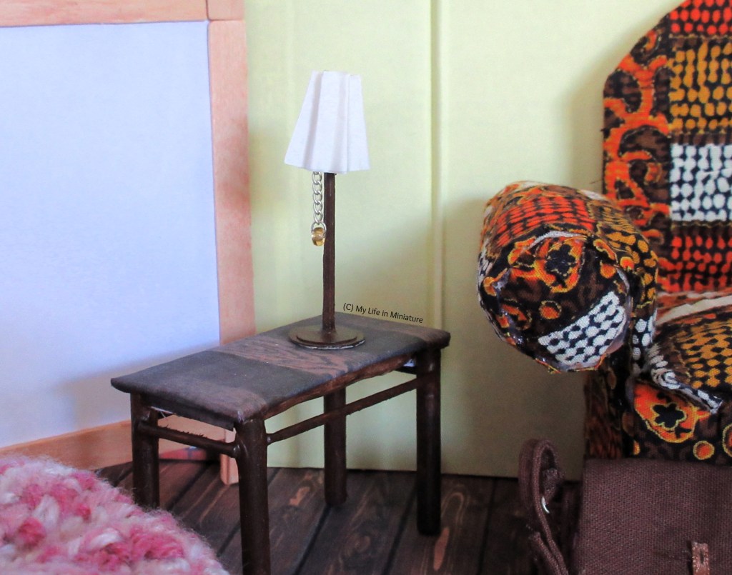 Close-up of the side table beside the armchair. The table has a dark wood surface, and painted dark wood legs. The small lamp on the table looks very similar to the freestanding lamp, with dark wood base and post, white lampshade, and silver chain hanging from beneath the shade. 
