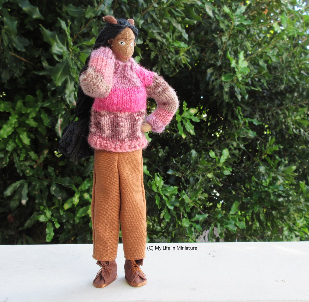 Petra stands against a large green bush, wearing the brown pants and her pink jumper. She has one hand behind her neck and the other on her hip, and is looking to the right. 