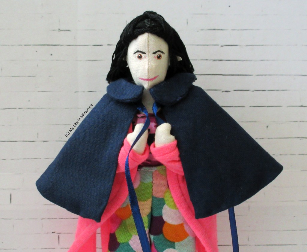 Tiffany stands against a white brick background. She wears colourful pants and a hot pink cardigan, and is pulling a navy cape over her shoulders. She is smiling at the camera.