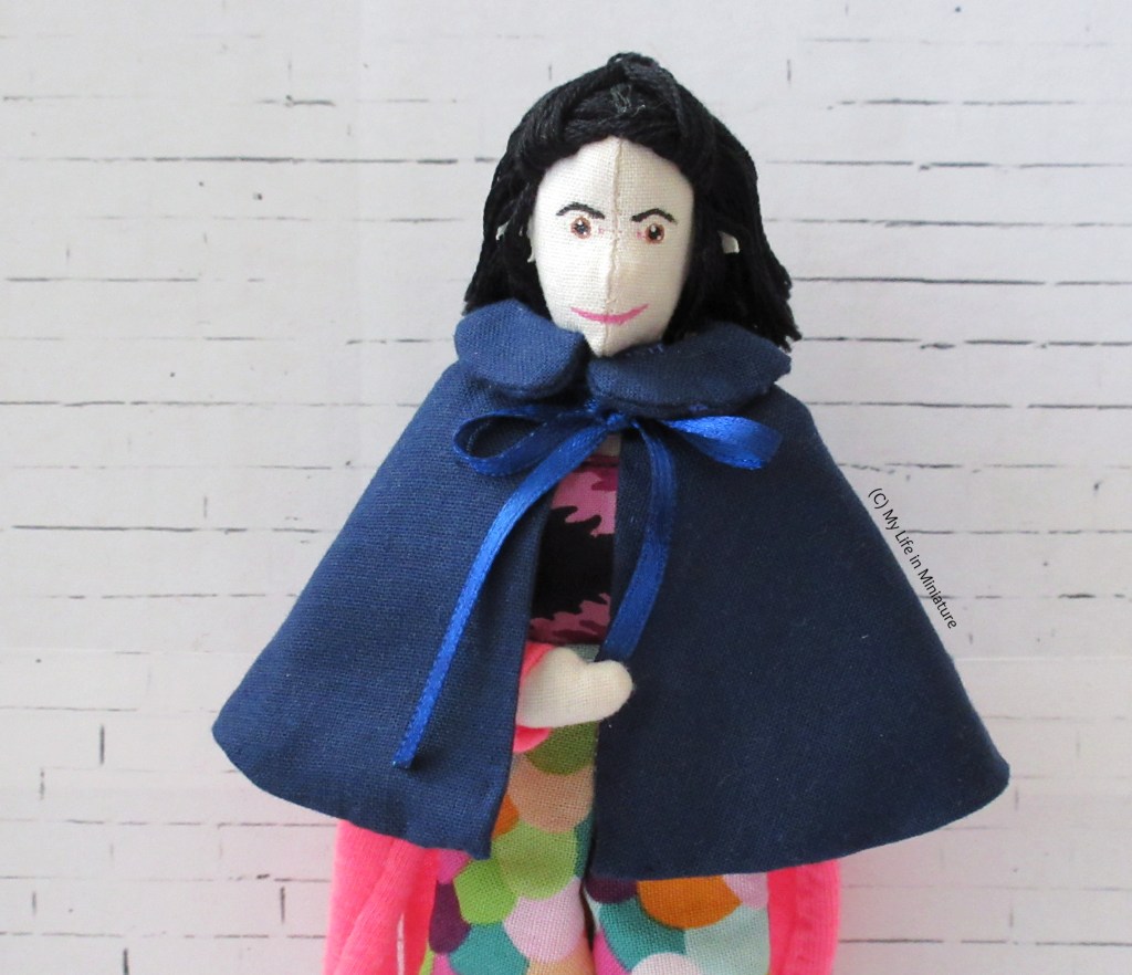 Tiffany stands against a white brick background. She has a navy cape tied around her shoulders. The cape is about wrist-length, with navy ribbon ties and a Peter Pan style collar.