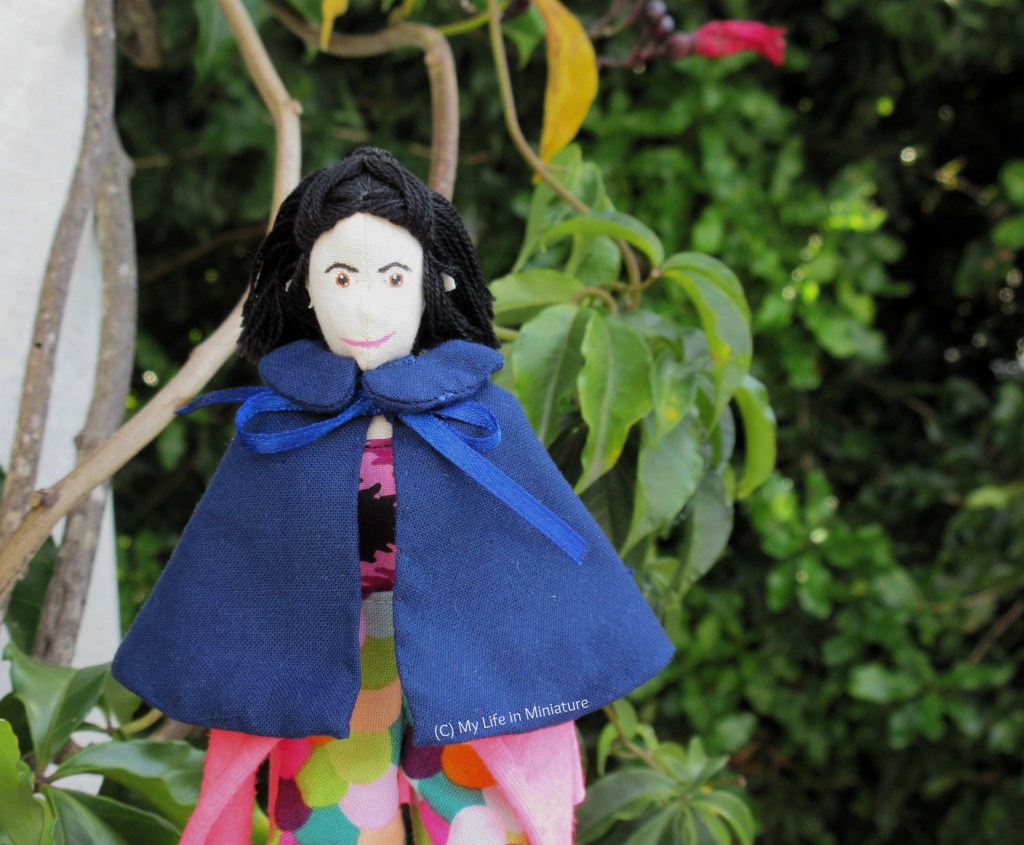 Tiffany stands near a white wooden post, which has a vine growing up it. She wears the navy cape, tied with a bow in the front. She has her hands in her pockets and is smiling at the camera. In the background is some leaves from the vine, and a large bush.