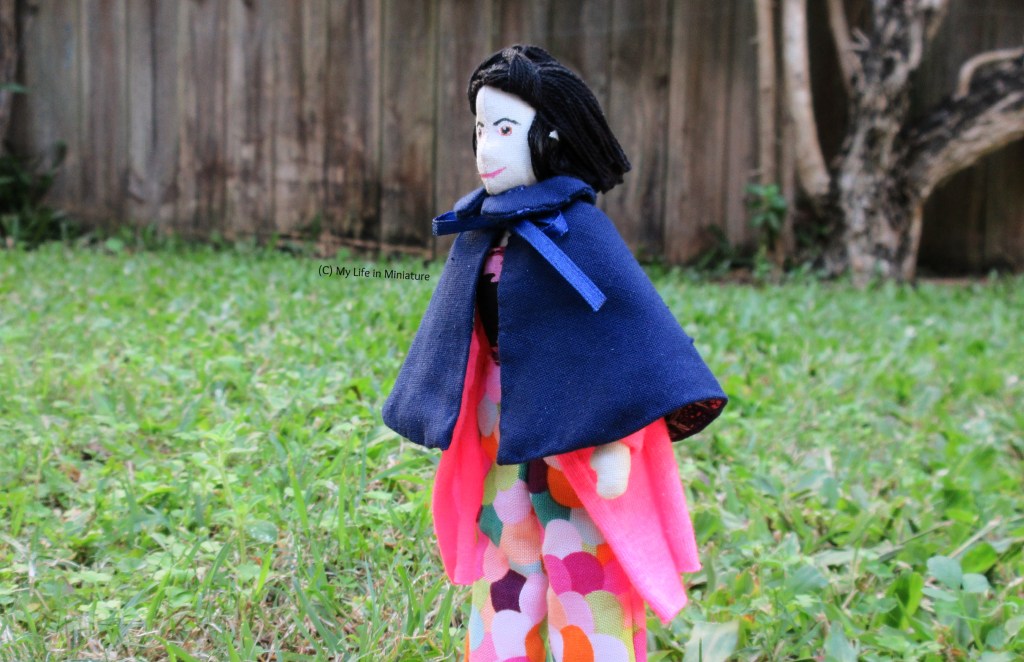 Tiffany stands in a 'field', and in the background is a wooden fence and a gnarled tree trunk. She wears the navy cape over her hot pink cardigan and colourful pants. Her body is angled to the left, which is also the direction she's looking.