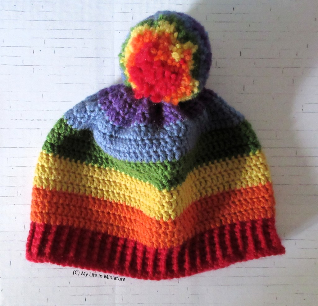 A rainbow-striped crocheted beanie with a rainbow-striped pompom sits flat-ish on a white brick background. The stripes are in rainbow order up from the ribbing.