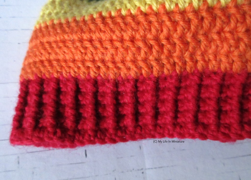 Close-up of the red ribbing of the rainbow-striped beanie on a white brick background. The treble crocheted orange stripe above the ribbing is also visible.