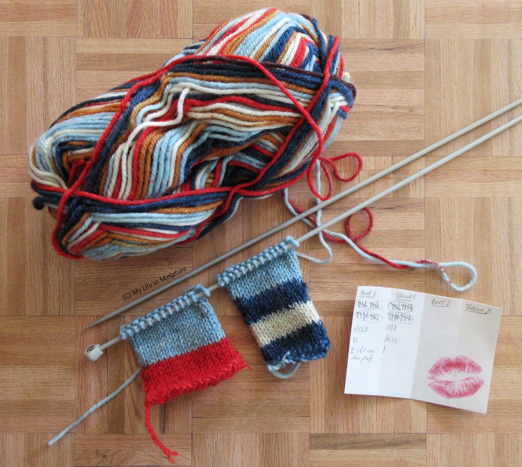 A ball of yarn, two knitting needles, two small pieces of knitting, and a sticky note sit on a wood background. The yarn is multicoloured: red, dark and light blue, white, and pale brown.