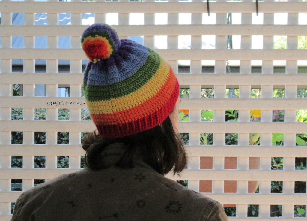 The author wears the rainbow-striped crocheted beanie, back to the camera with her head slightly turned to the right. Behind the author is a white lattice, and she has shoulder-length dark hair and a khaki jacket on.