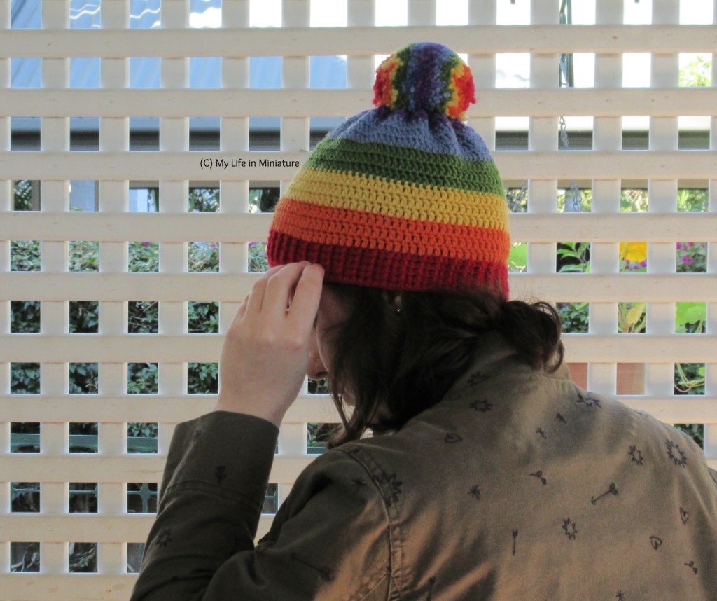 The author wears the rainbow beanie against a white lattice. Her head is turned almost fully to the left, but her face is blocked by her hand, which reaches up to adjust the ribbing.
