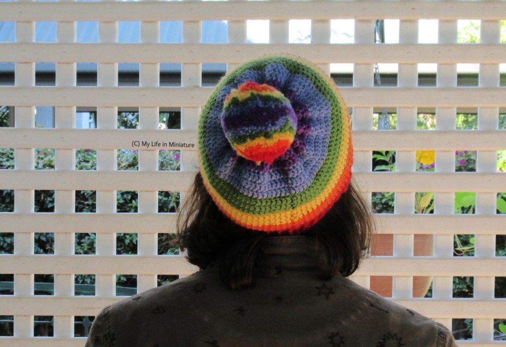 The author wears the rainbow beanie, back to the camera and against a white lattice. Her head is tilted back so the rainbow-striped pompom is visible from the top of the beanie. 