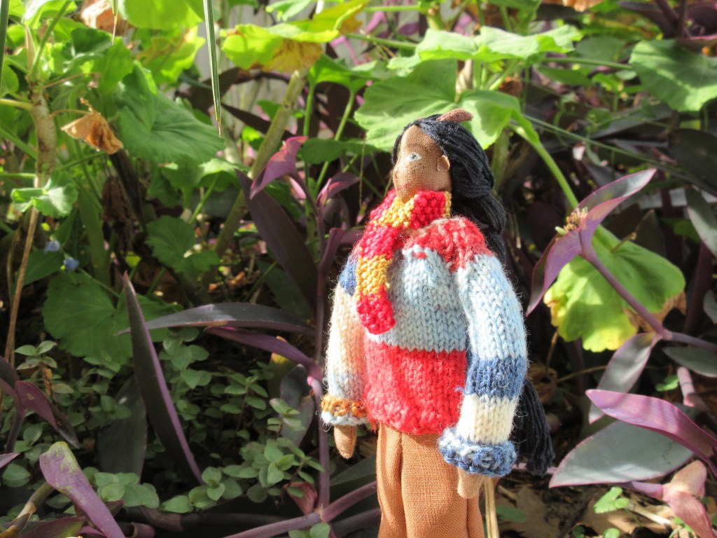 Petra stands outside in amongst leafy plants in green and purple. She looks up at the plants, face half-shaded by a leaf. She wears her multi-coloured jumper with the sleeves turned up, brown pants, and a red-and-gold knitted scarf. 