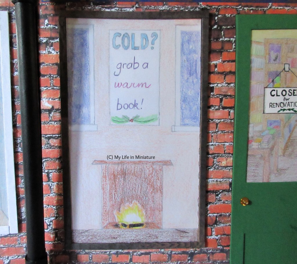 The front window of The Palace Library is visible. A wintery display is in the window, depicting a small scene around a fireplace. Above the fireplace (in which a fire burns) is a piece of 'art' that says 'Cold? Grab a warm book!'. Part of a window to a dark blue snowy exterior is visible on each side of the artwork.