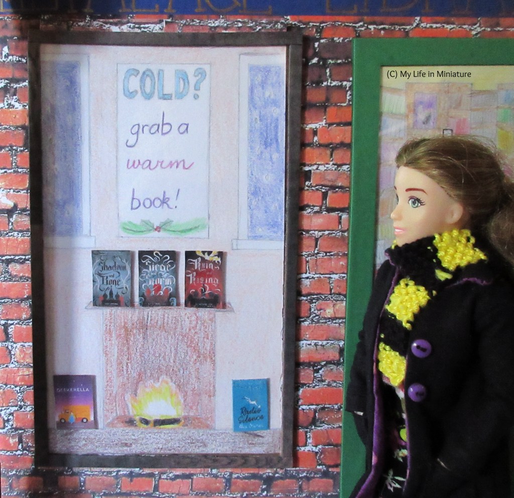 Sarah stands beside The Palace Library's front window, looking at it. The window has the wintery display with a fireplace and the text 'Cold? Grab a warm book!' above it. Three books sit on the mantlepiece of the fireplace, and two more are on the floor on either side of the fire.