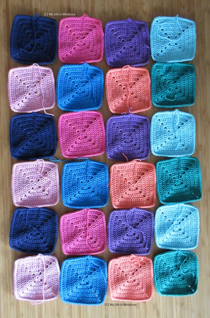 All 24 crocheted squares are laid out in a long rectangle, four by six squares. They are laid out in colour order - navy, pink, purple, pale blue, pale pink, blue, peach, and green.