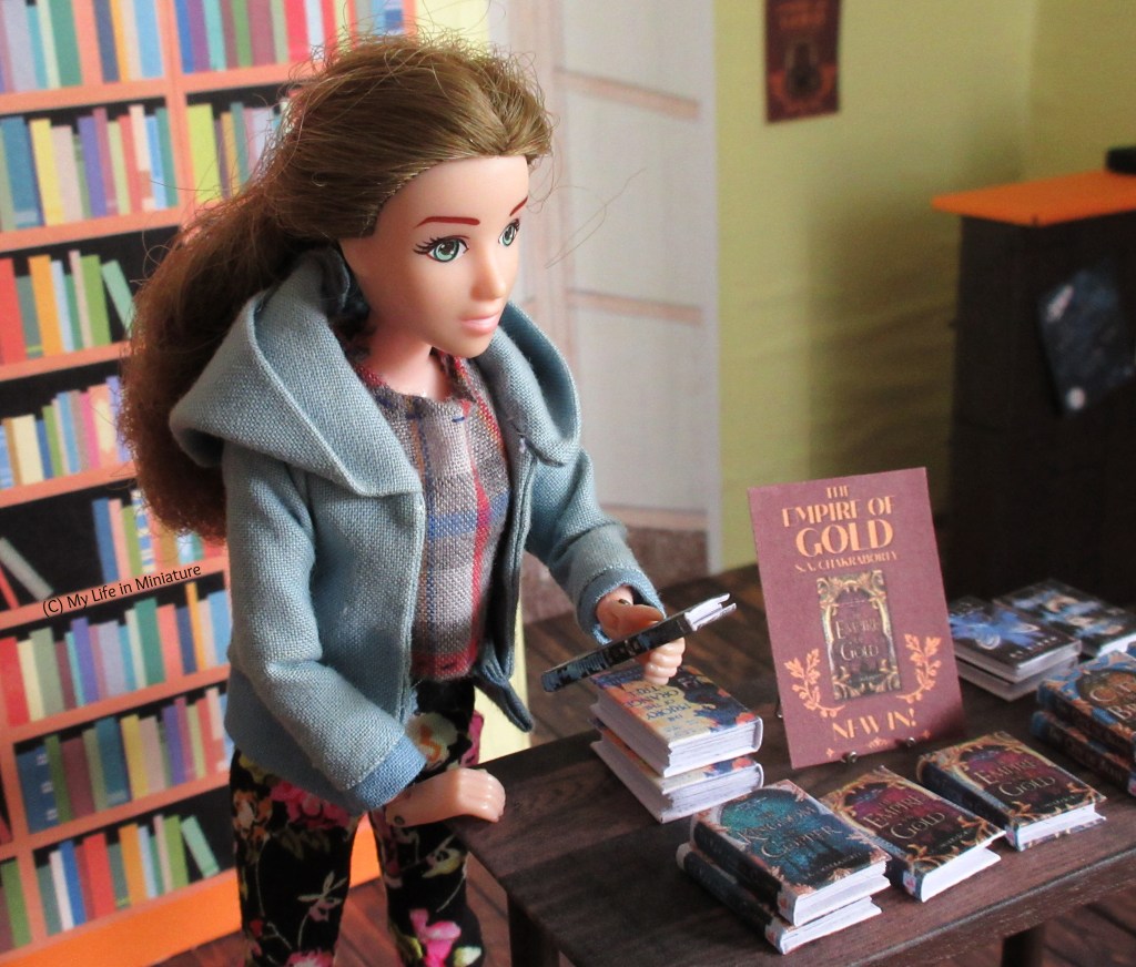 Sarah stands at a corner of the free-standing table in the Palace Library, wearing a blue hoodie and floral pants. She is looking at a book she's holding, called 'The Weight of the Stars', reading the blurb with interest.