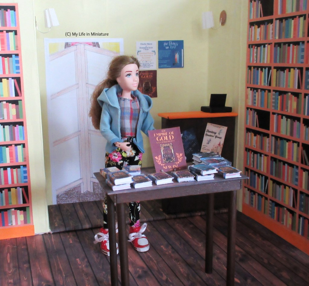 Sarah stands in the centre of the counter area, display table in front of her. She gestures around to the space, and looks to the right. The display table has several stacks of books on it, with a flyer advertising one of them on a wire book stand. A counter is in the background, as well as a doorway blocked by a room divider and bookshelves.