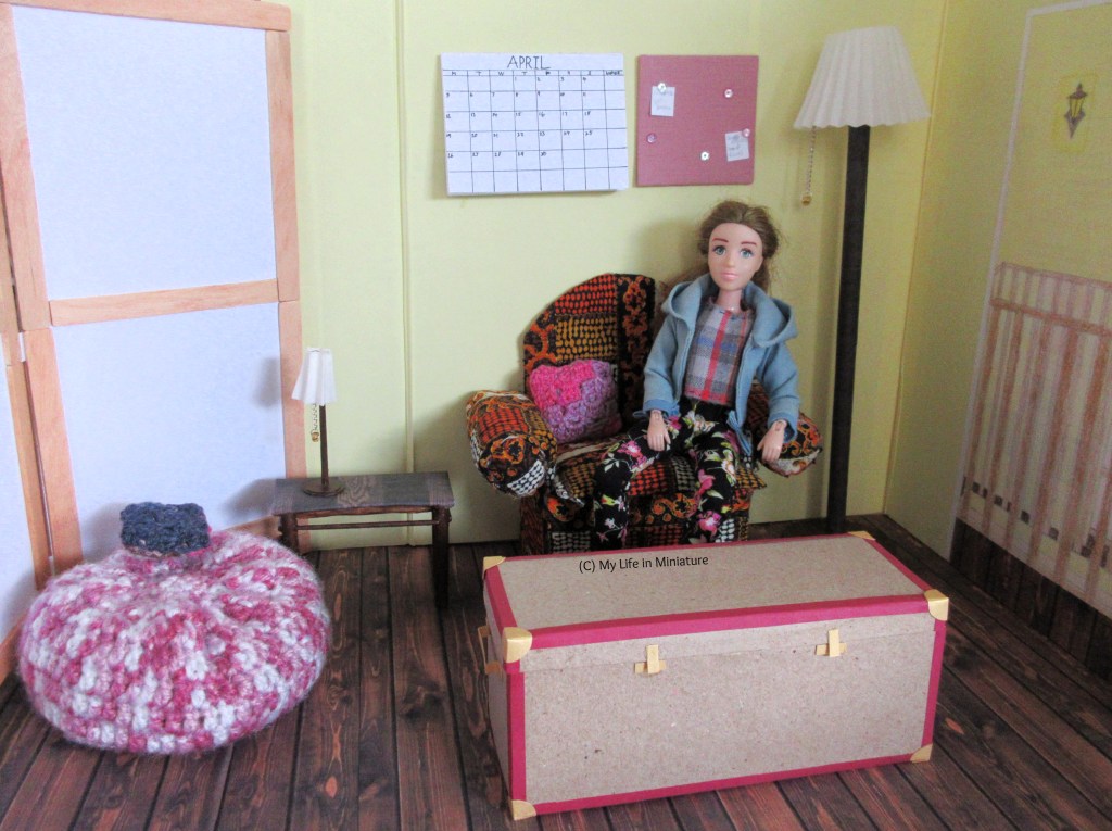 Sarah sits in an armchair in an upstairs room of the Palace Library. To the right of the room is the doorway, with a floor lamp beside it. In front of the armchair is an old chest, and beside it is a small side table with a lamp on. In the left-hand corner is a beanbag with a blue cushion on. A calendar, set to April, is on the wall behind the armchair, next to a noticeboard.