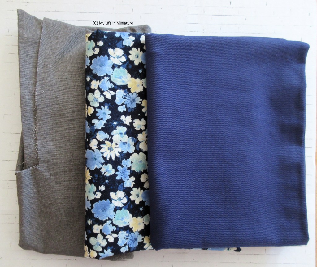 Three folded-up pieces of fabric sit on a white brick background. They are stacked in a staggered manner, so they are all visible. Left-right, there is a grey cotton that has clearly been used, a dark blue cotton printed with pale blue, white, and yellow flowers, and a heavier dark blue fabric. 
