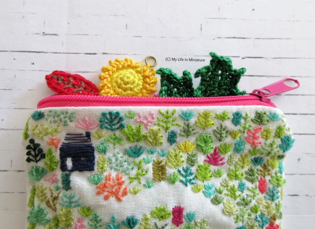 An embroidered zipper pouch sits, open, on a white brick background. Peeking out the opening at the top of the bag is a small crocheted red leaf, sun, and green leaves. 