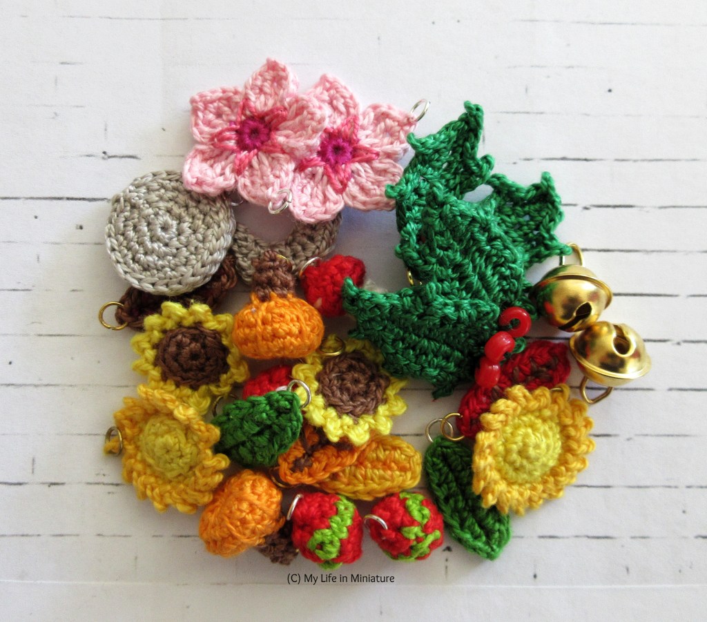 A pile of small crocheted objects is on a white brick background. The objects all have a jump ring on them, and are mostly crocheted natural objects - apart from two gold bells on the right of the pile.