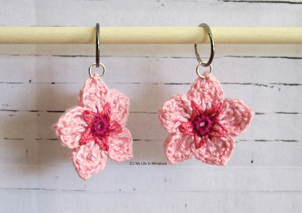 The two crocheted cherry blossom flowers hang by their silver jump rings from silver sleeper earrings. The silver sleepers are on a wooden dowel, against a white brick background.