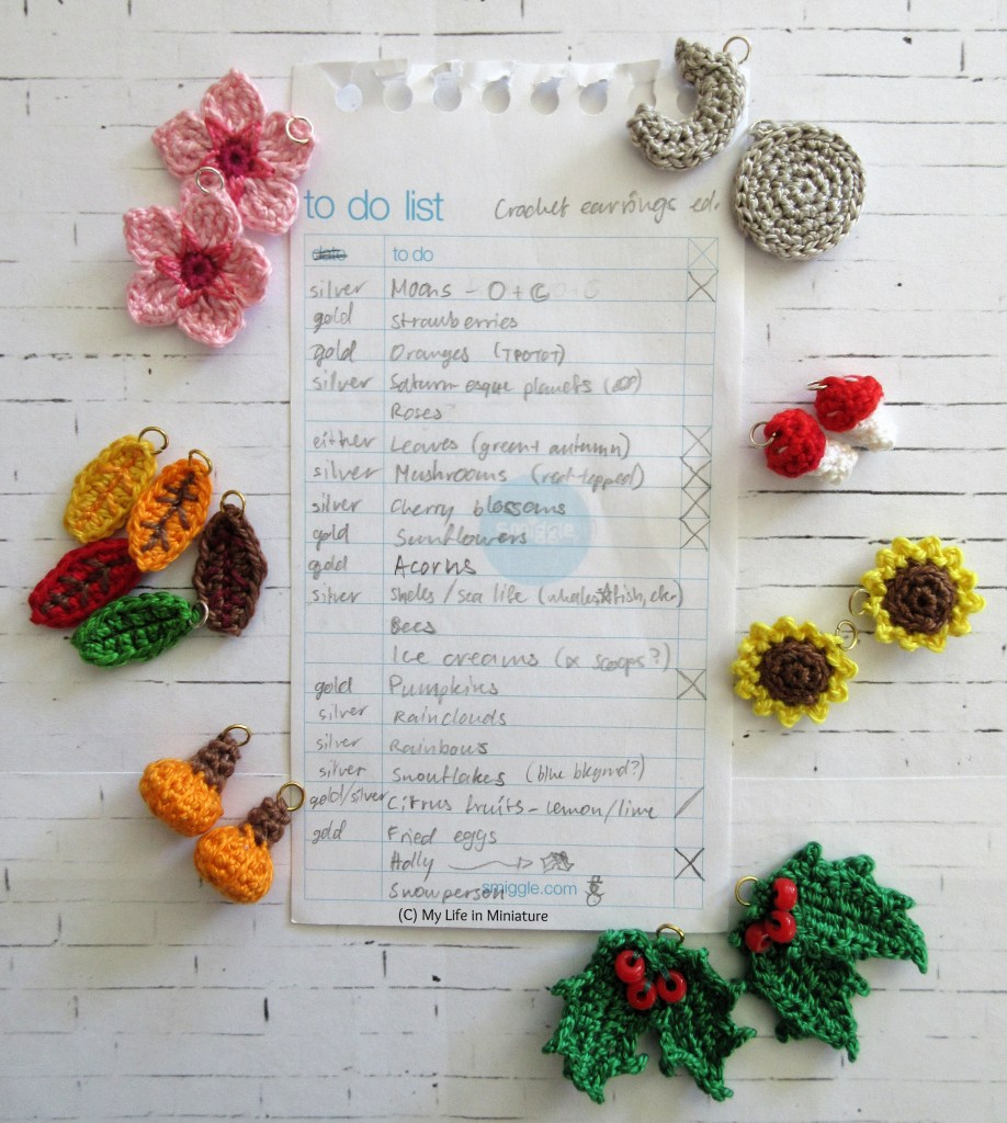 A hand-written list is surrounded by various small crocheted objects, on a white brick background. The list is titled 'To Do List: Crochet Earrings Edition'. Several of the items are crossed off, and the completed earrings sit roughly beside the list. The completed items include sunflowers, mushrooms, moons, cherry blossoms, autumn leaves, pumpkins, and holly.