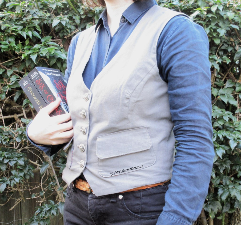 The author stands in front of a hedge, wearing a grey waistcoat, a navy button-up shirt, and black jeans. She carries the Six of Crows duology by Leigh Bardugo. She is white with shoulder-length brown hair, just visible on the collar of the shirt.