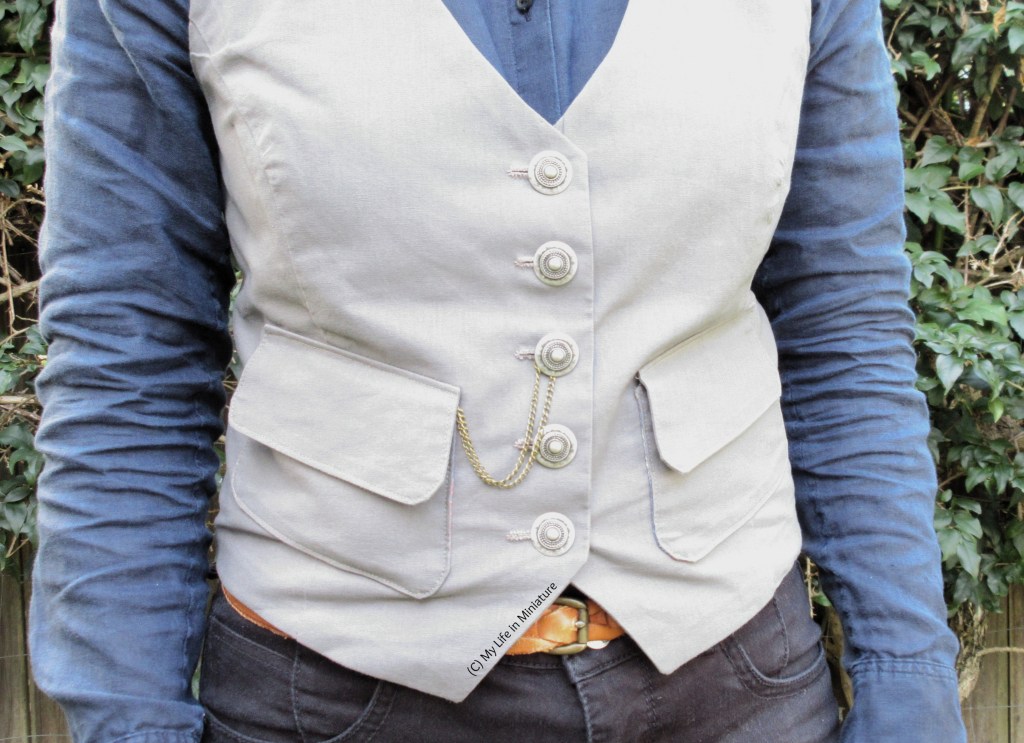 The author stands in front of a hedge, wearing a grey waistcoat, a navy button-up shirt, and black jeans. From the left pocket emerges a chain, which loops over the centre button and hangs loosely. 