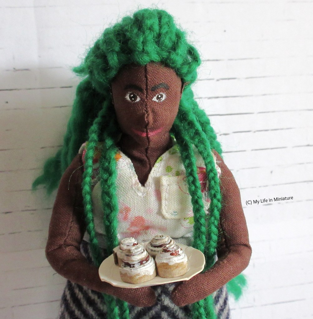 Hazel stands in front of a white brick background, holding a plate of four cinnamon rolls. She looks down at the plate. She has some braids pulled forwards over her shoulders, and wears a white v-necked sleeveless top with pink flowers on.