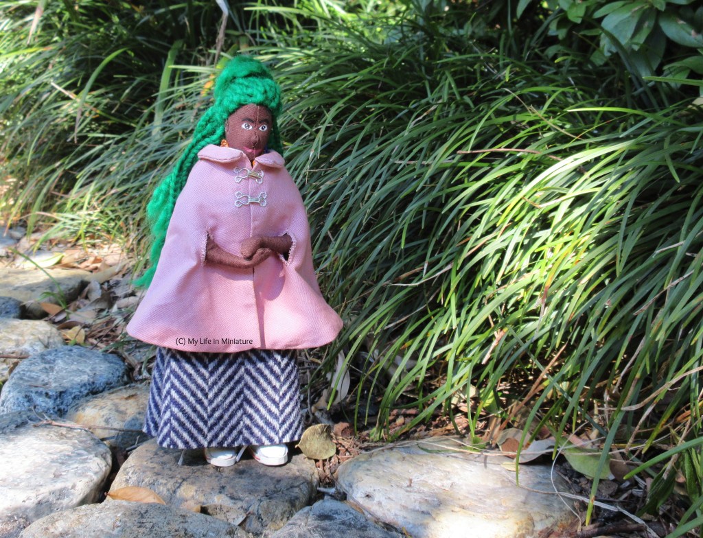 Hazel walks next to some long, thin, bushy plants on a path laid with circular stones. She wears her dusky pink cape and her wool skirt with white shoes. Her hands are clasped in front, and she looks down at where she's stepping. 