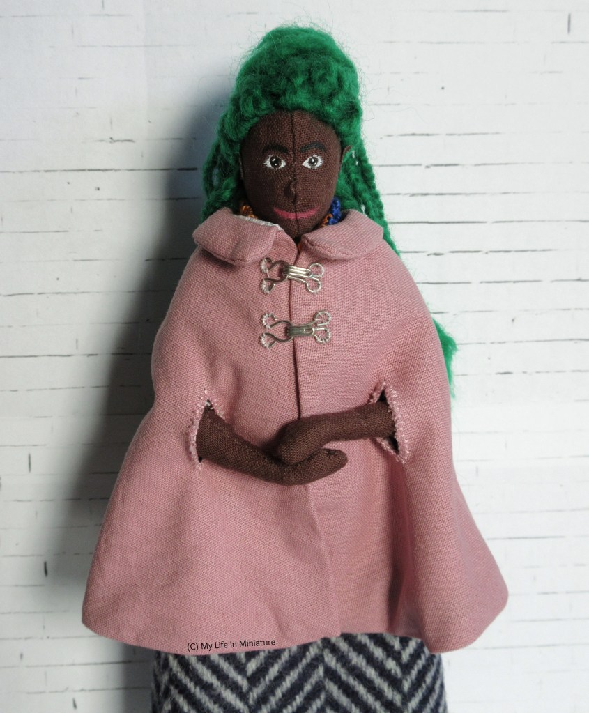 Hazel stands against a white brick background. She wears a dusky pink knee-length cape with a zig-zagged grey-and-navy wool skirt. The cape has a Peter Pan collar, two armhole slits, and attaches at the chest with two silver hooks-and-eyes. Hazel has her hands clasped and smiles at the camera.