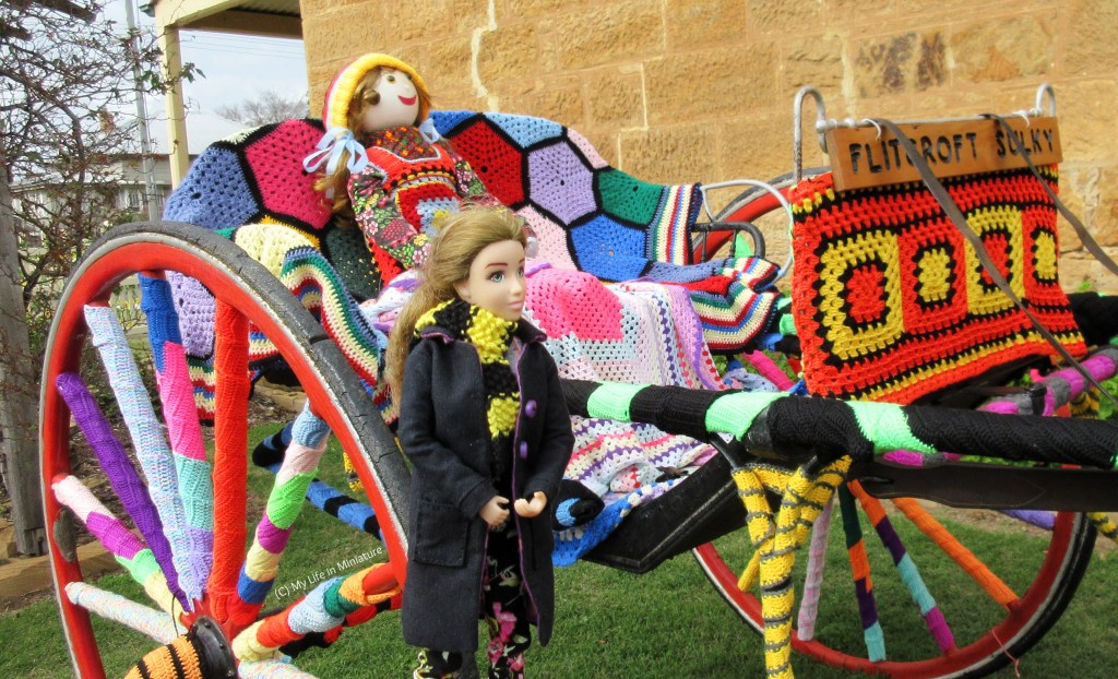 A different angle of the crochet-covered small carriage with the horse hitched to the front. The horse is out of shot, and the seat of the carriage is more visible. Sarah stands in front of it, looking to the right as if looking into the seat of the carriage. 