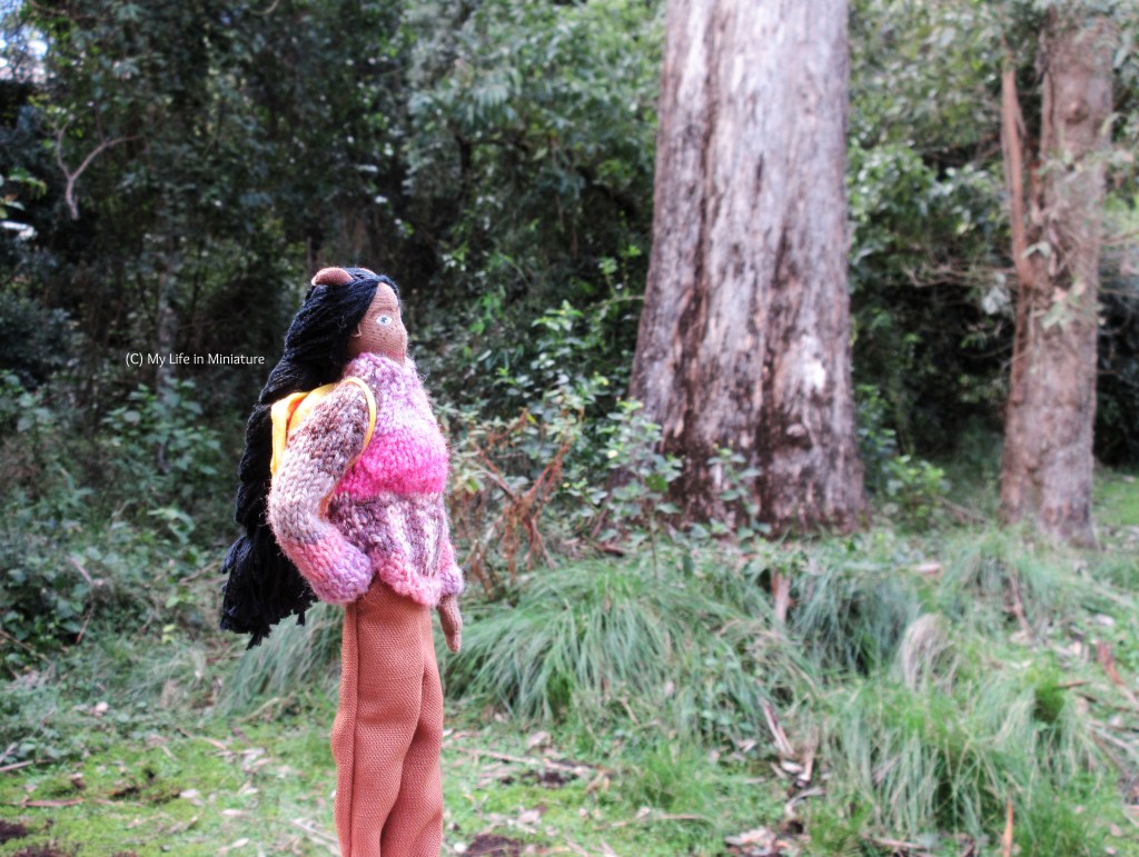 Petra stands near the base of a tree trunk, facing right, head tipped back to try and see the treetop. She wears a pink-and-purple jumper with brown pants, and has her yellow backpack on. She has her closest arm in her pant pocket. 