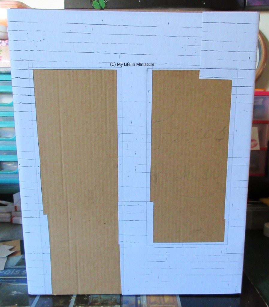 The front of a cereal box fills the image, covered in cardboard that peeks out from behind white brick paper. Two roughly rectangular holes are left in the paper for a door and a window, which the cardboard is visible through.