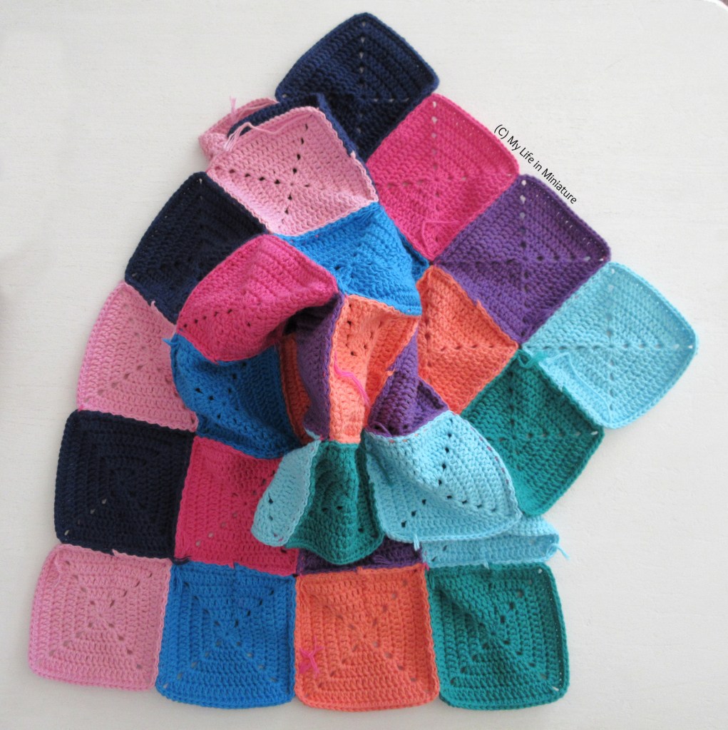 32 crocheted squares, of various colours, are sewn together to make a long rectangle of fabric. This rectangle is curled and scrunched up in the centre, so the entire length of the rectangle is not visible. It is on a white background. 