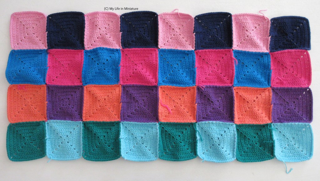 32 crocheted squares, of various colours, are sewn together to make a long rectangle of fabric. It sits on a white background. A few loose ends are visible around the edges of the rectangle.