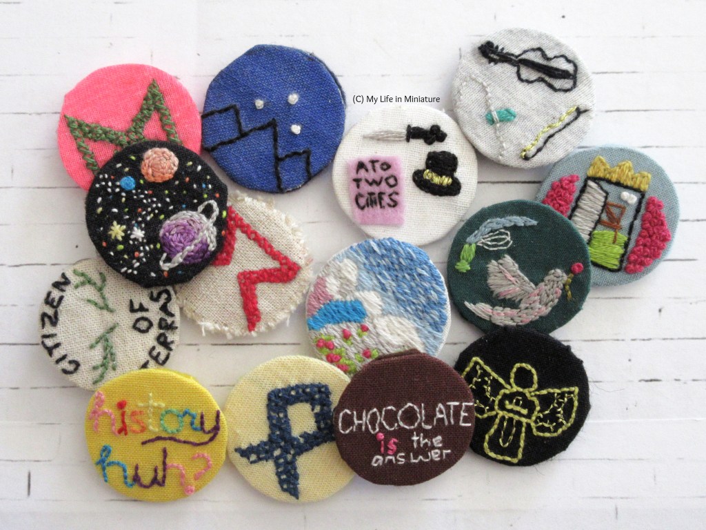 About 15 embroidered pin badges are in a loose pile on a white background. Most of them relate to books like the Infernal Devices series, the Magnus Chase series, and Red, White and Royal Blue. At the bottom, the 'Chocolate is the answer' badge can be seen.