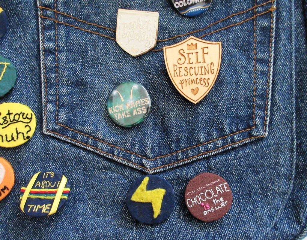 Image shows the front of a denim satchel bag. The bag has a number of pin badges on it, with one being the embroidered 'Chocolate is the answer' badge. Others include a wooden shield that says 'Self Rescuing Princess', and a white glittery pocket-shaped one that says 'Pocket Equality'. 