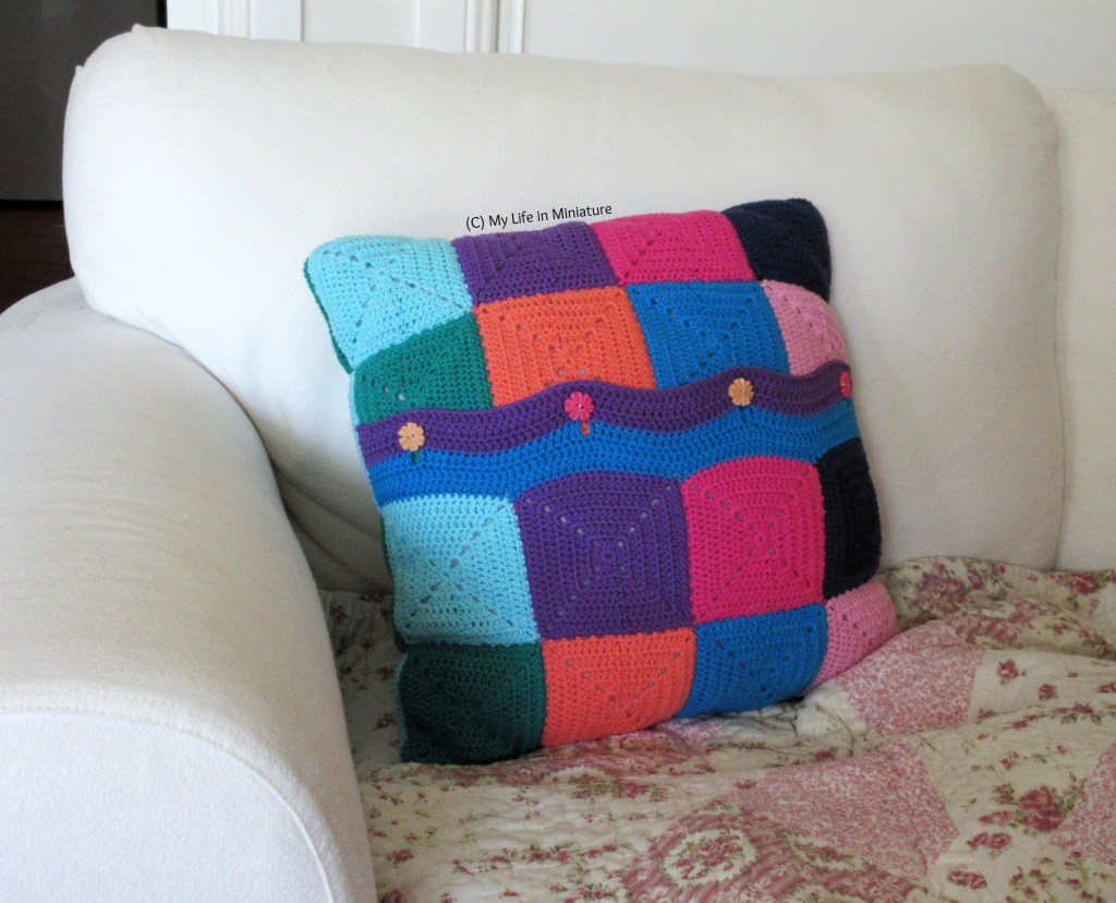 A crocheted cushion cover sits on a white couch. It is made of crocheted squares in various bright colours, with four buttons buttoned into a crocheted-on strip that covers the opening.